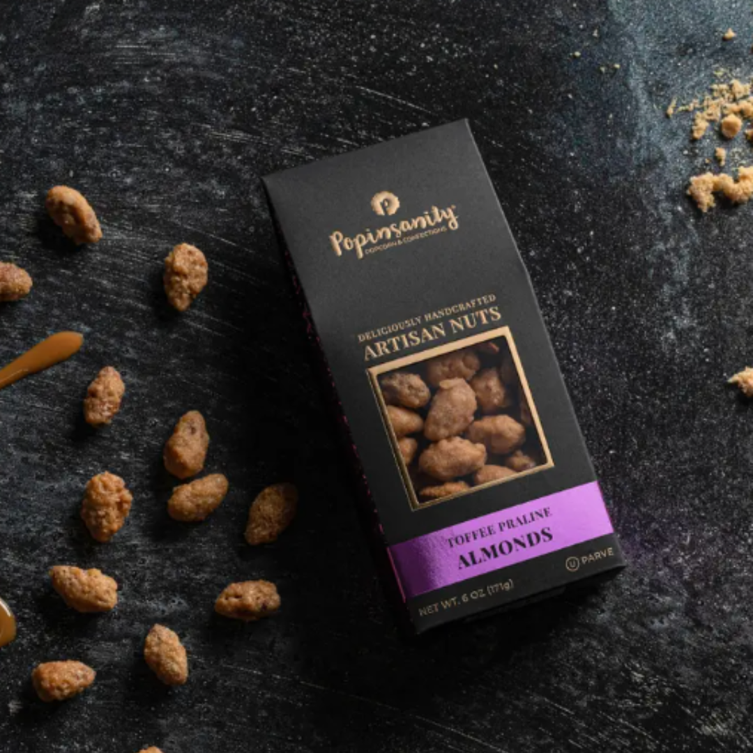 Beautiful black package of Handcrafted Toffee Praline Almonds with No Artificial Flavors.  The box of almonds is laying on marble table with a few almonds scattered around the package.