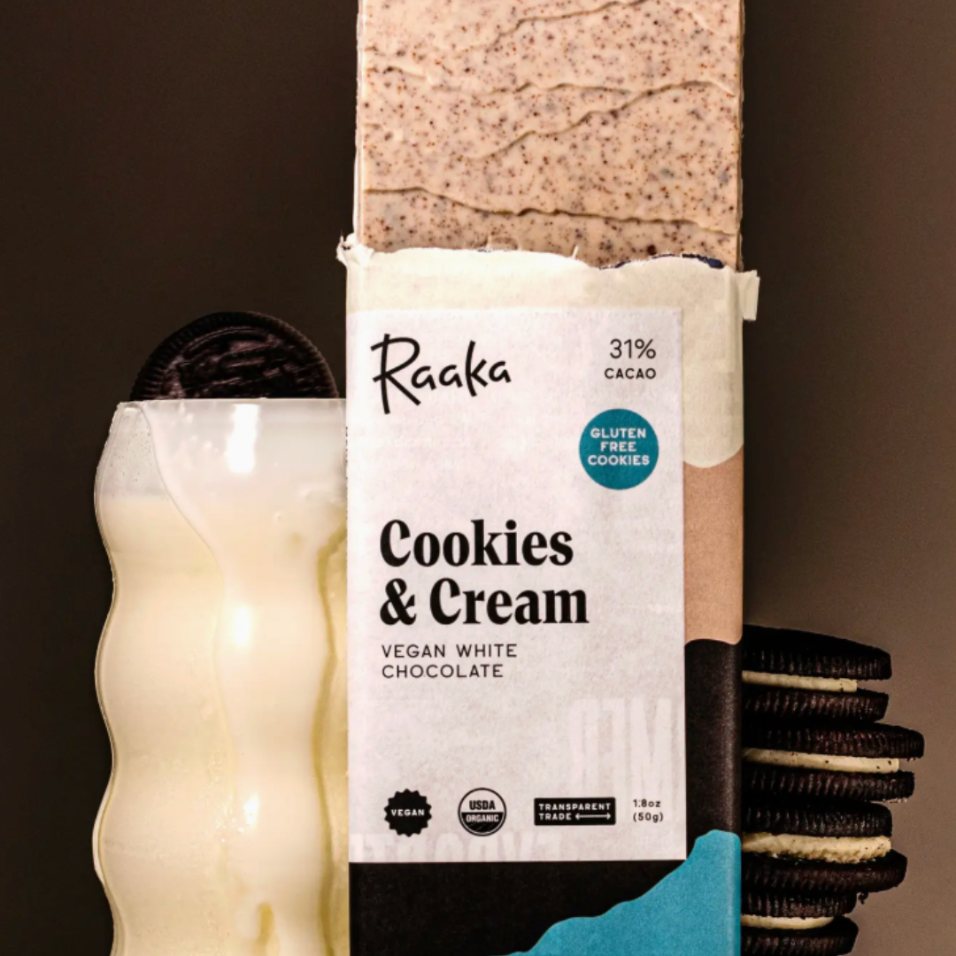 Raaka Cookies & Cream vegan white chocolate bar with packaging on a dark background with a glass of milk.