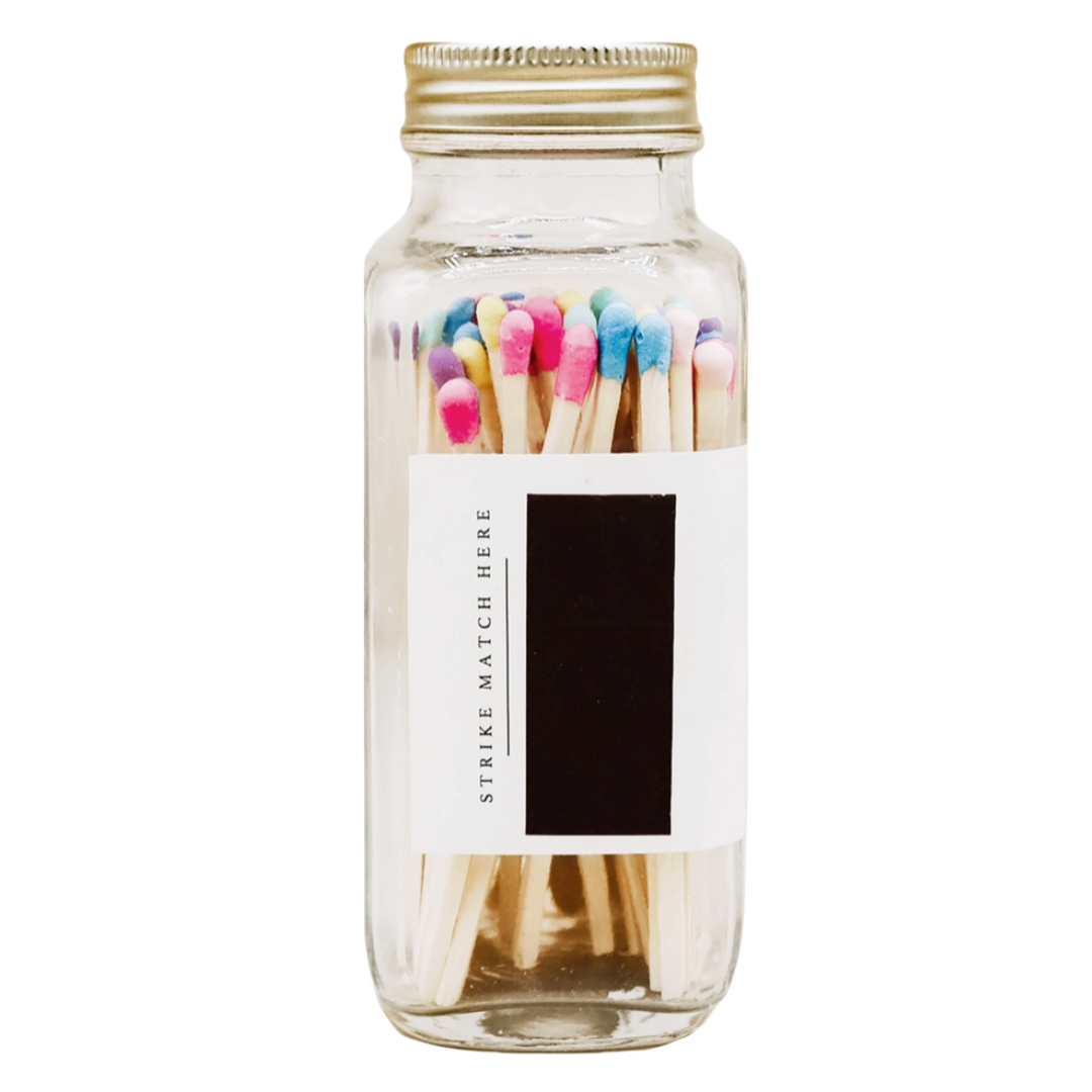 Glass bottle filled with wooden matches with pink, blue, purple, yellow, peach tips and a gold lid against a white background. Features the strike pad on the side of the bottle.