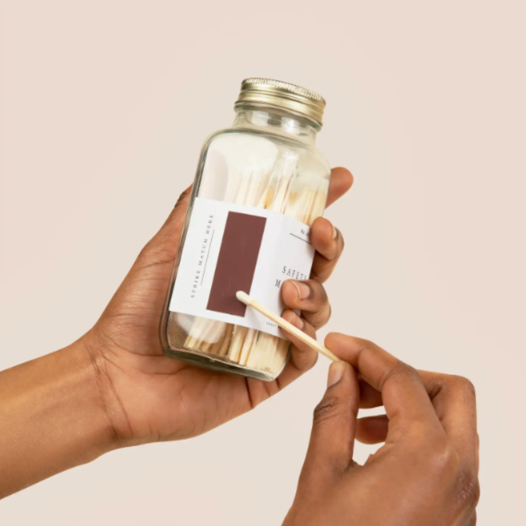 Person holding a jar of matches with a matchstick, against a plain background