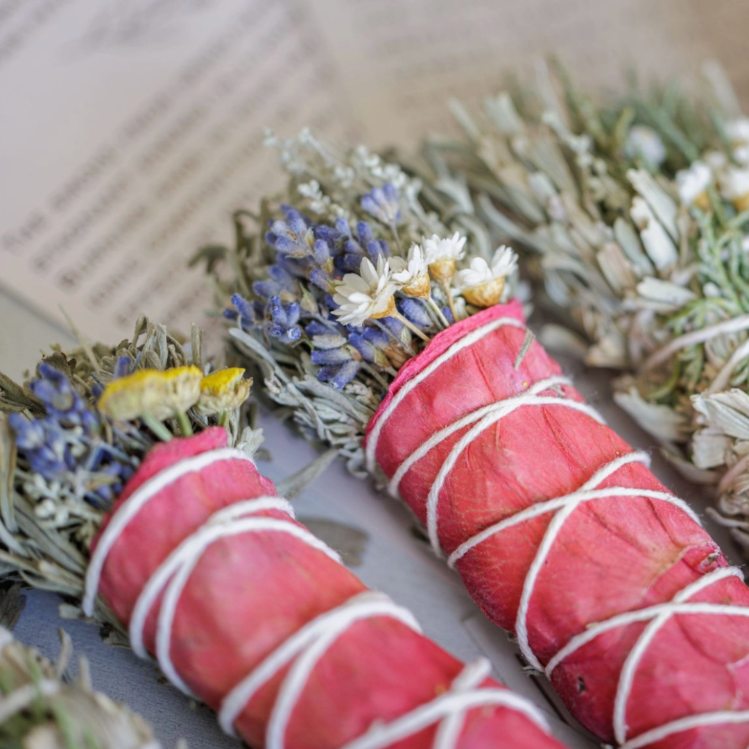 A close-up of a meticulously crafted Floral Sage Bundle, featuring sagebrush and various herbs and flowers, with a crystal (amethyst, clear quartz, or citrine) attached. The bundle is presented on a soft, neutral surface.