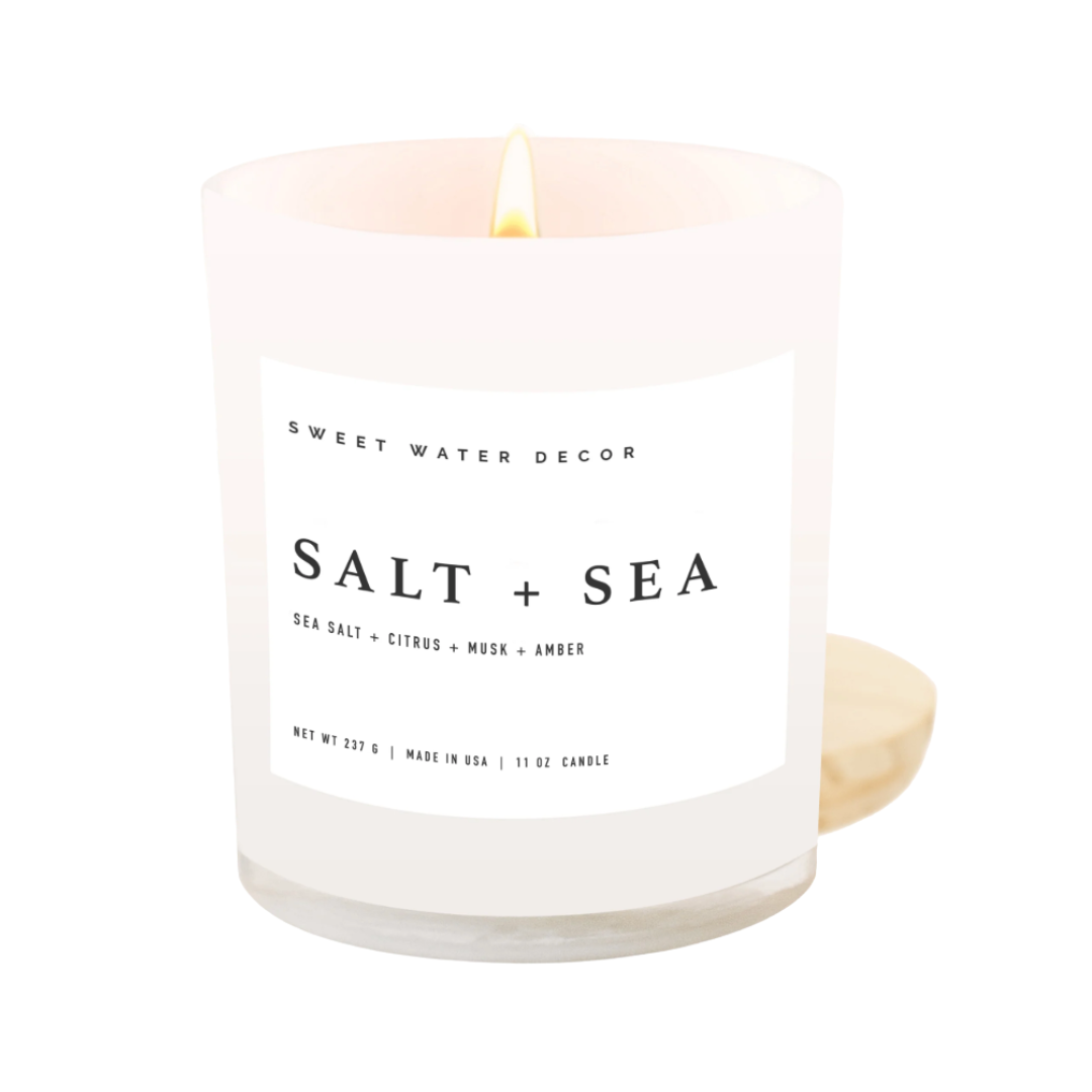 White glass candle labeled 'Salt + Sea' by Sweet Water Decor on a white background. The candle has a wooden lid.