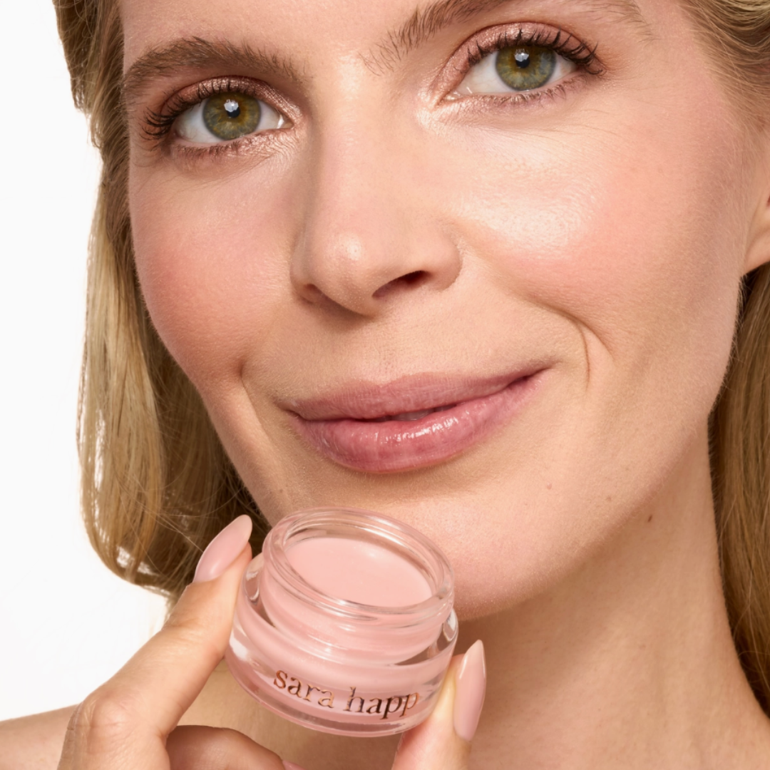Woman holding a pink lip balm container with 'sara happ' branding.