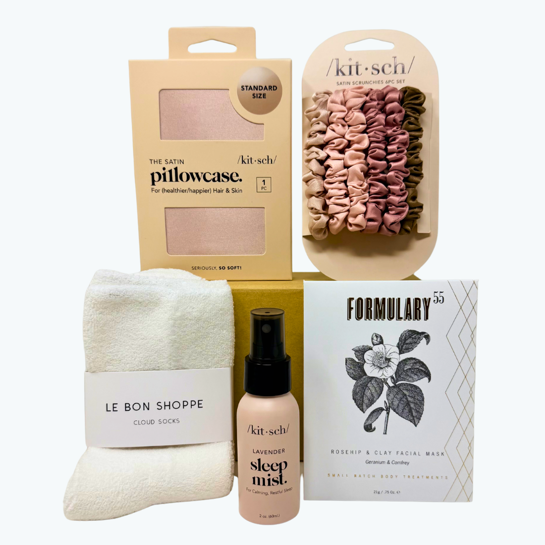 Collection of beauty and lifestyle products including a pink satin pillowcase, lavender sleep mist, set of six pink satin hair scrunchies and facial mask packaging on a white background.