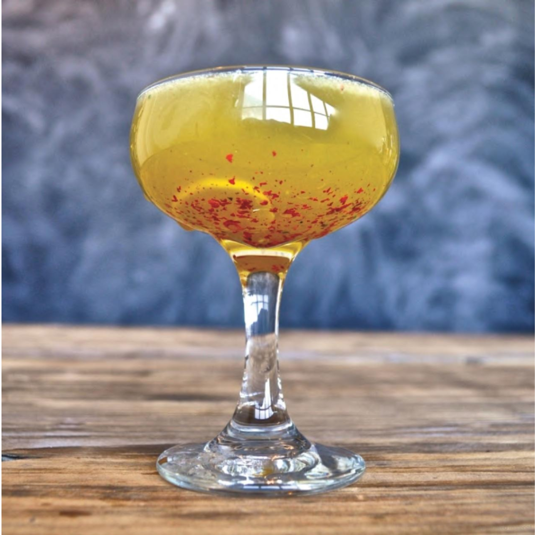 Shake Cocktail Book featuring seasonal recipes for simple and fun cocktails. A martini glass is displayed on a wooden table with a yellow cocktail with speckled red ingredient floating towards the bottom of the glass.