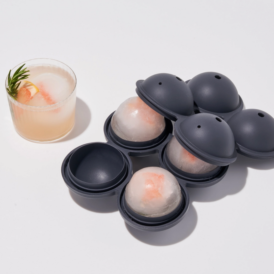 Ice ball mold with ice balls and a cocktail on a white background. The silicone ice mold is grey and makes four spheres.