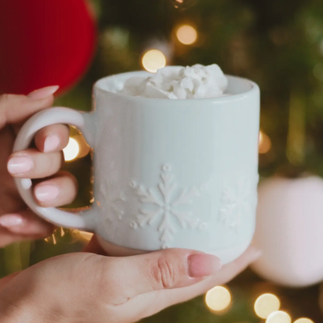 Person holding a light blue mug with hot chocolate and whipped cream against a festive background.