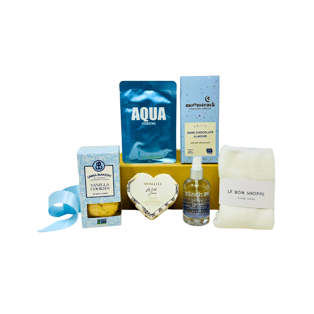 Collection of blue and white products in a gift box including sugar cookies, chocolate bar, cozy socks, heart shaped body buffer, aqua facial sheet mask and lavender chamomile pillow spray.