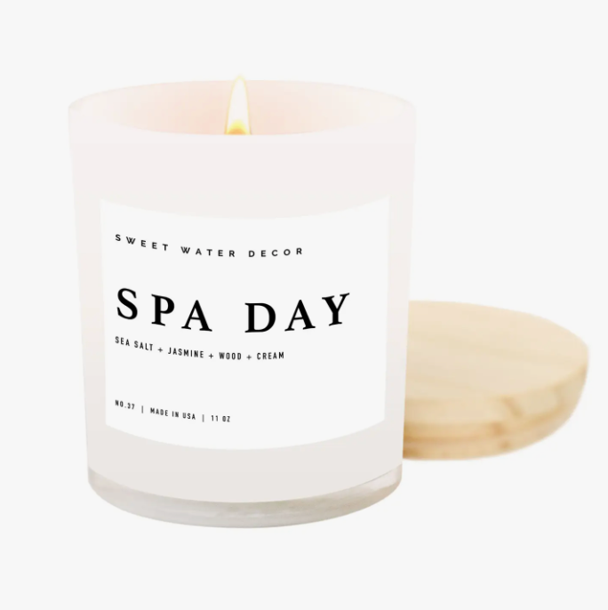 'Spa Day' Soy Candle: A white glass jar candle with a wooden lid, offering a tranquil ambiance for self-care rituals. This 11oz candle features a soothing blend of sea salt, jasmine, woody, and creamy notes, with a burn time of over 50 hours. Crafted with a cotton wick and soy-based wax, it's vegan, non-GMO, and free from phthalates and toxins.