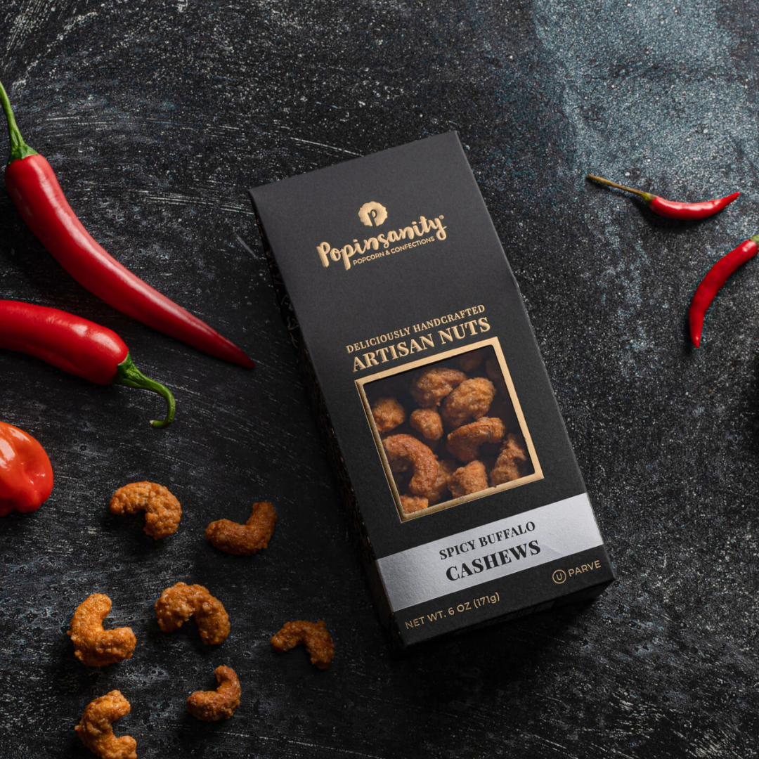 Box of Popin' Sanity spicy buffalo cashews on a dark surface with cashews and red peppers.