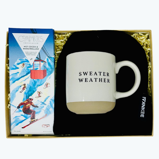 Gift set with a mug labeled 'Sweater Weather', a beanie, and a hot chocolate bar in a decorative box.