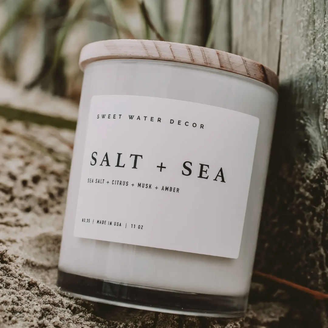 Candle labeled 'Salt + Sea' by Sweet Water Decor on a sandy background.