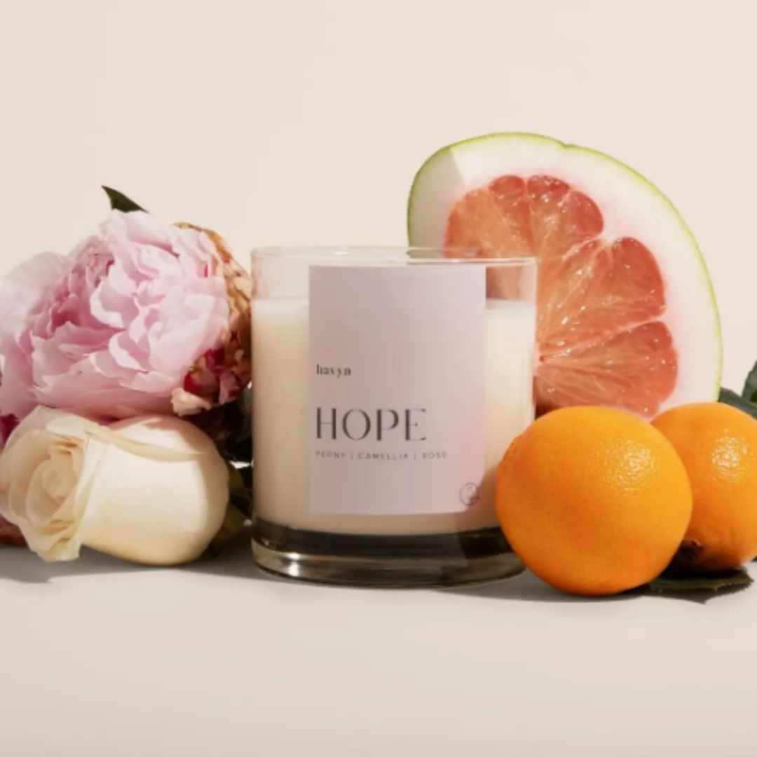 The HOPE Signature Candle in a clear glass jar, showcasing its minimalist design with "HOPE" engraved on the front. The candle, made of a coconut and soy wax blend, has a calming scent combination of Peony, Camellia, Rose, Pomelo, and Mandarin. It is hand-poured and intended to create a relaxing atmosphere, with a burn time of 40 hours and a fragrance compliant with EU standards.