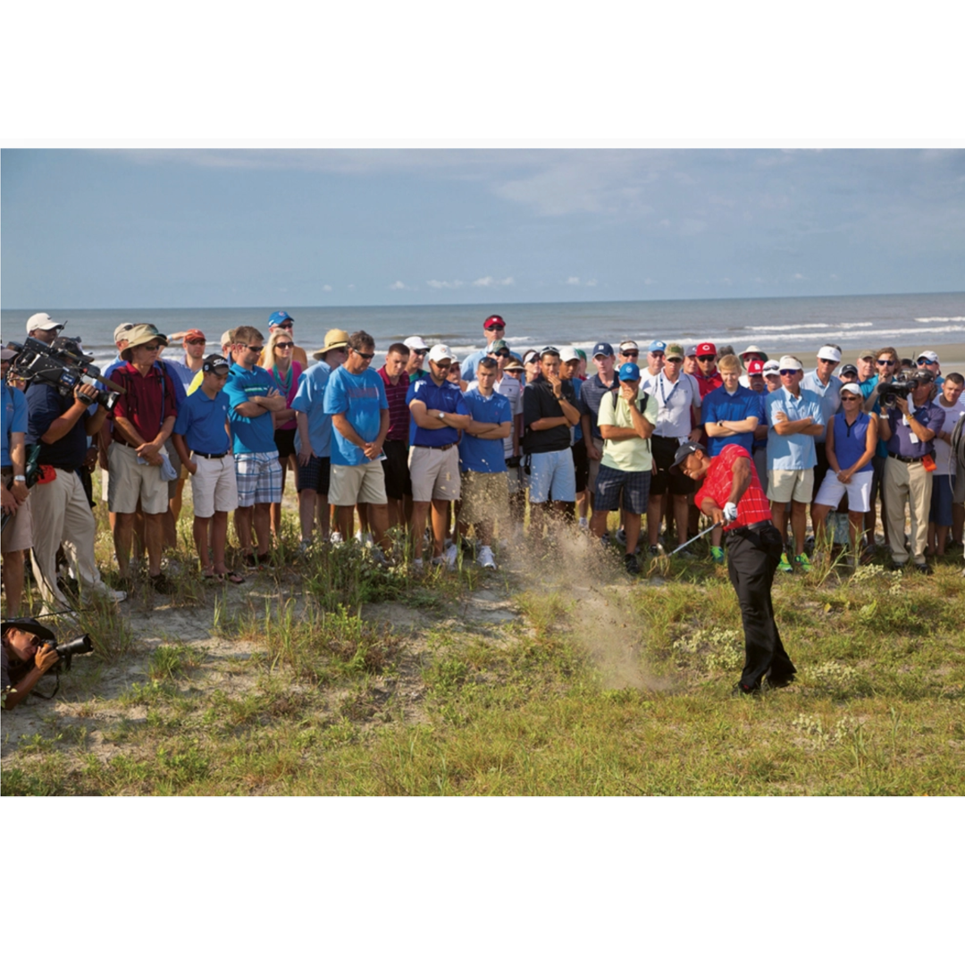 Golfer swinging a club on a grassy area with a crowd watching, near a body of water.
