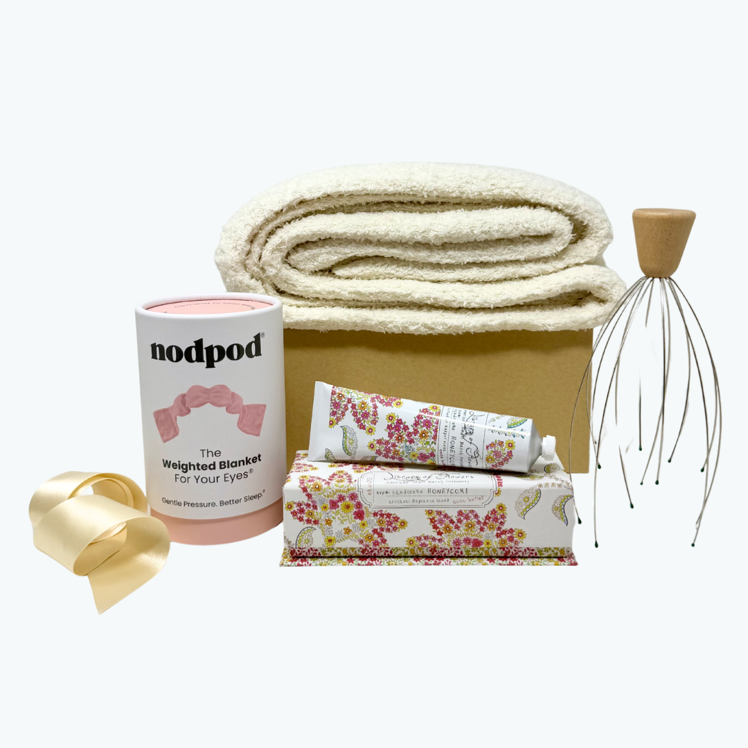 Unwind Wellness Kit Gift Box with cream blanket, blush weighted sleep mask, hand cream in a pink floral gift box and a head massager with wooden handle on a white background.
