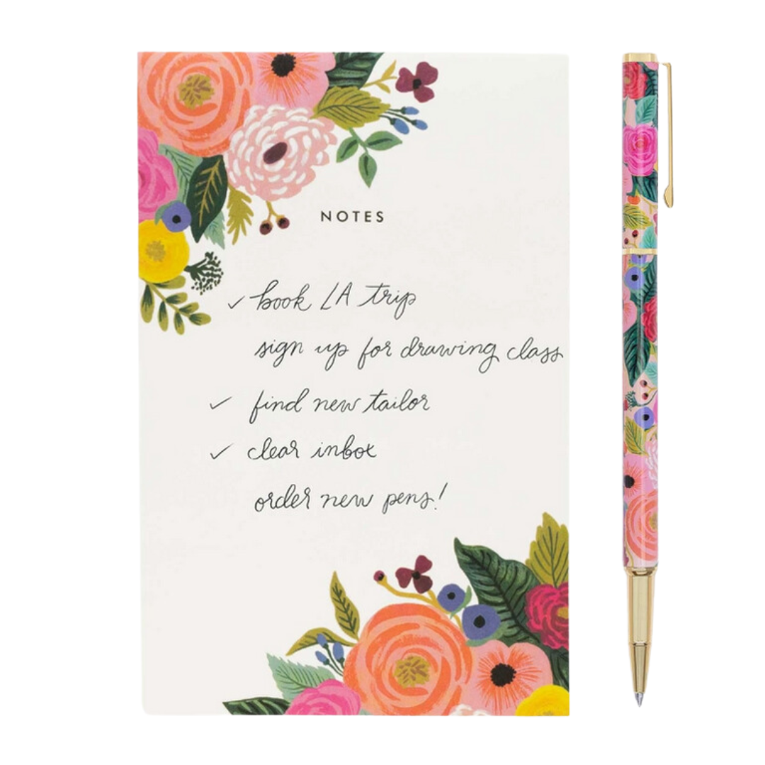 Close-up of the Garden Party Pen by Rifle Paper Co. next to a floral piece of note paper. The pen’s stainless steel body and brass details are visible, showcasing its 0.5 mm rollerball tip and vibrant floral design.