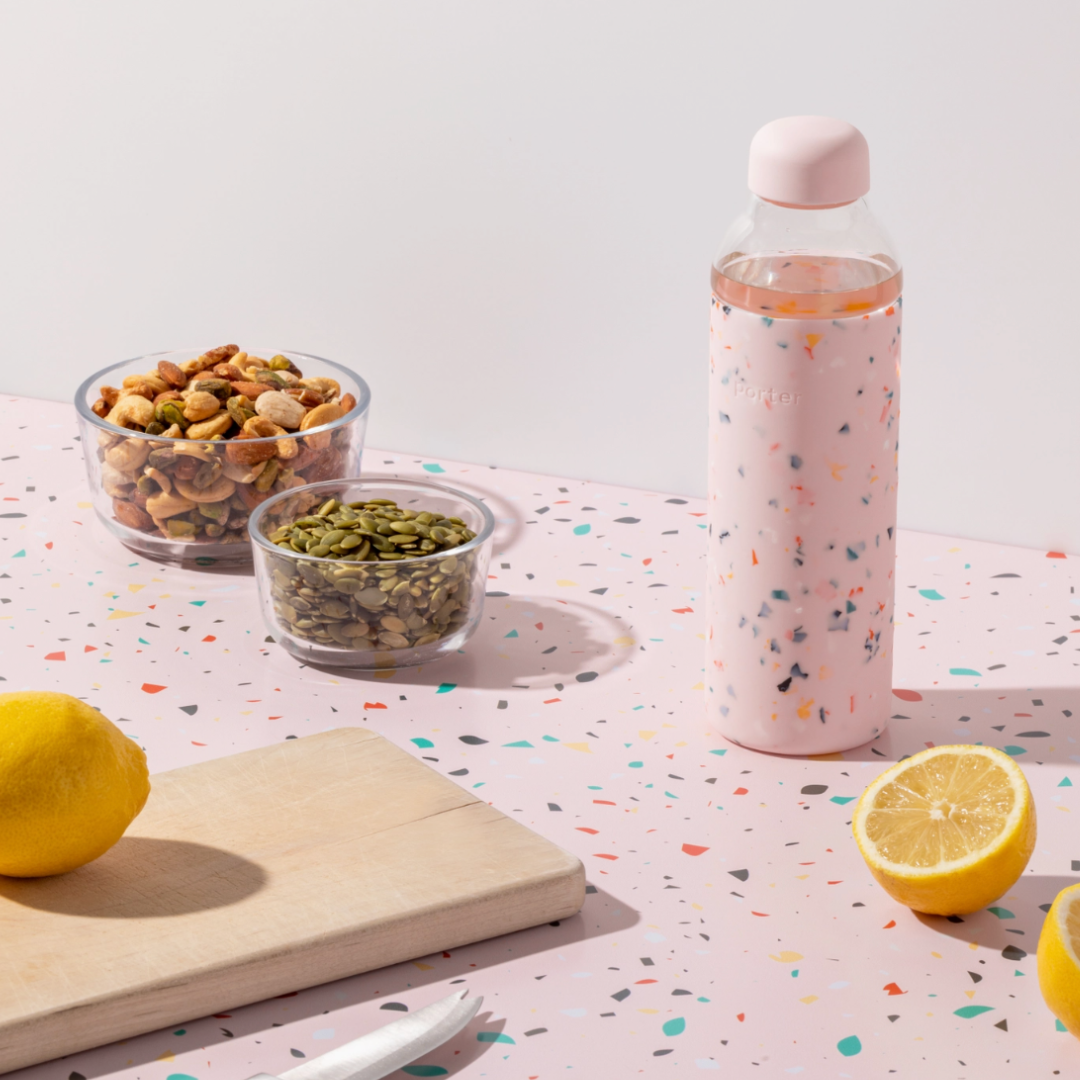 Porter's 20oz glass and pink silicone reusable water bottle shown on a table next to bowls of nuts and seeds.