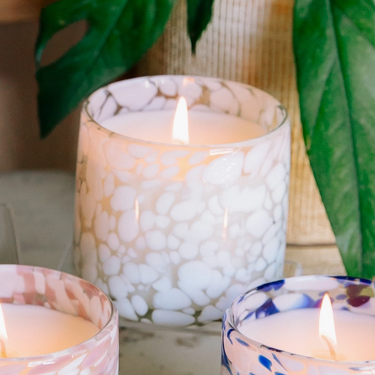 Three lit candles in decorative confetti glass holders with a blurred green leaf background.