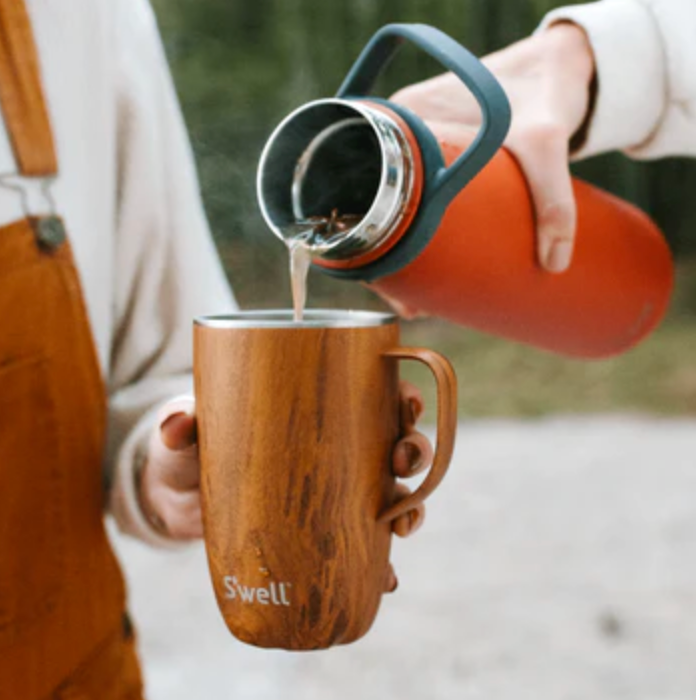 A 16oz stainless steel mug featuring a warm teak-wood finish and a sturdy handle and sliding lid, perfect for enjoying your favorite drinks on the go. The tumbler is being held in someone's hand while someone else is filling it with a liquid beverage in an outdoor setting.