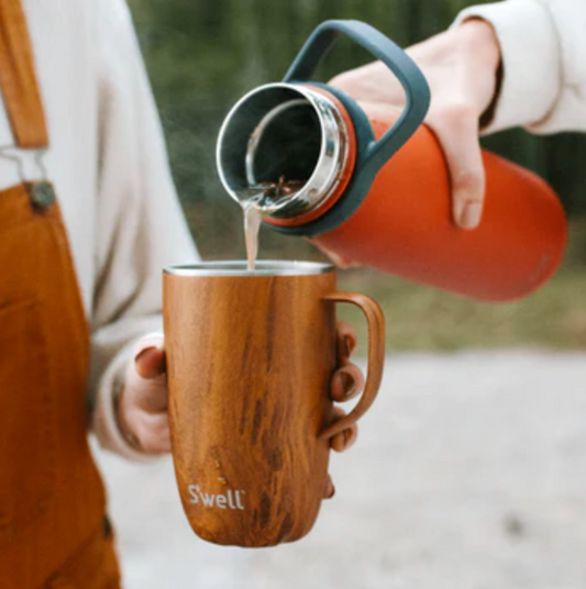 A 16oz stainless steel mug featuring a warm teak-wood finish and a sturdy handle and sliding lid, perfect for enjoying your favorite drinks on the go. The tumbler is being held in someone's hand while someone else is filling it with a liquid beverage in an outdoor setting.