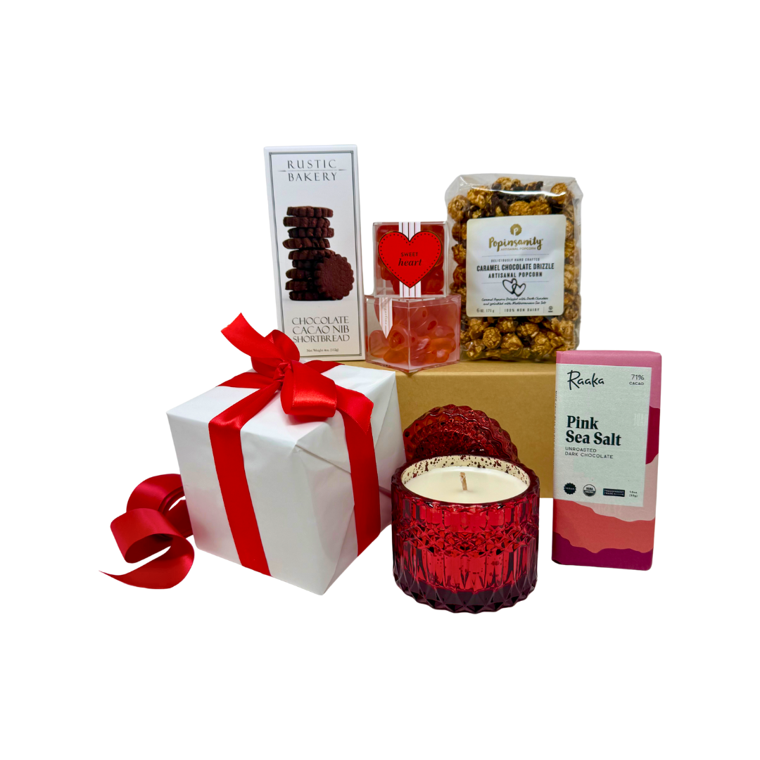 A curated gift box with a red glass shimmer candle, organic dark chocolate bar, chocolate shortbread cookies, strawberry gummy hearts in an acrylic box and a clear bag of caramel chocolate drizzle artisanal popcorn. The gift is wrapped in white paper with a red satin bow.